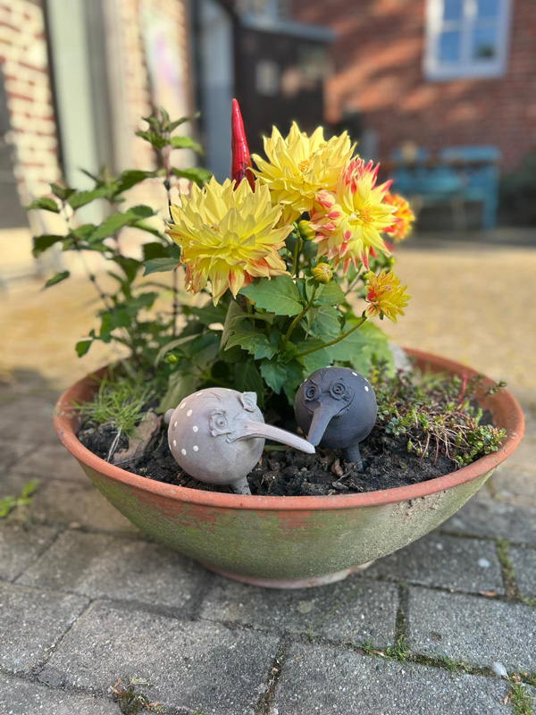 Garten-Keramik-Skulpturen In einer getöpferten und grünlich glasierten Schale sind gelbe Blumen und Minze eingepflanzt. In der Erde stehen zwei Kugelförmige Skulpturen, die an Vögelchen erinnern.