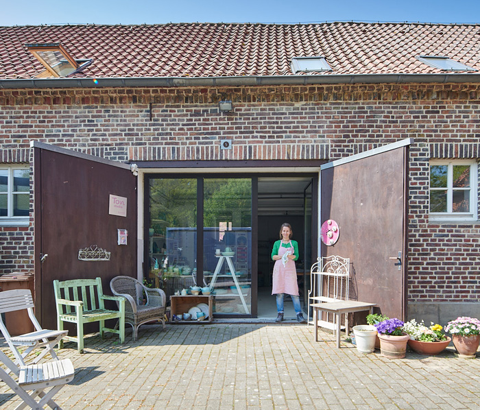 Ton-Studio-Keramik-Haus-Coerde Keramikkurs mit Kerstin Habersack bei einem Jugsellinnenabschied. Kerstin erklärt einer jungen Frau mit kurzen Bobhaarschnitt und grüner Schürze etwas zu Keramiken.