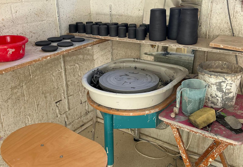 topferscheibe-ton-ecke getöpferte Keramiken aus Münster in grünlich-türkisen Glasuren stehen auf einem Tisch und mit Bäumen im Hintergrund. Sie werden von schönem Sonnenlicht gestriffen.